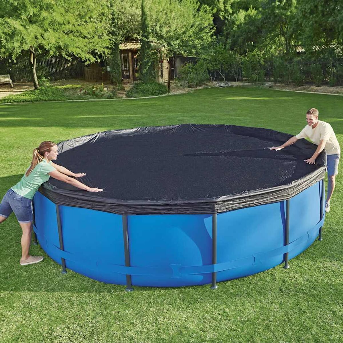 Zwei Personen bedecken ein blaues oberirdisches Schwimmbecken mit einer schwarzen Abdeckung auf grünem Gras.