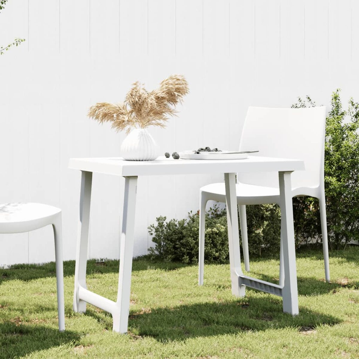 Weißer Tisch und Stühle im Freien auf Gras. Eine Vase mit getrockneten Pflanzen und ein weißer Zaun im Hintergrund.