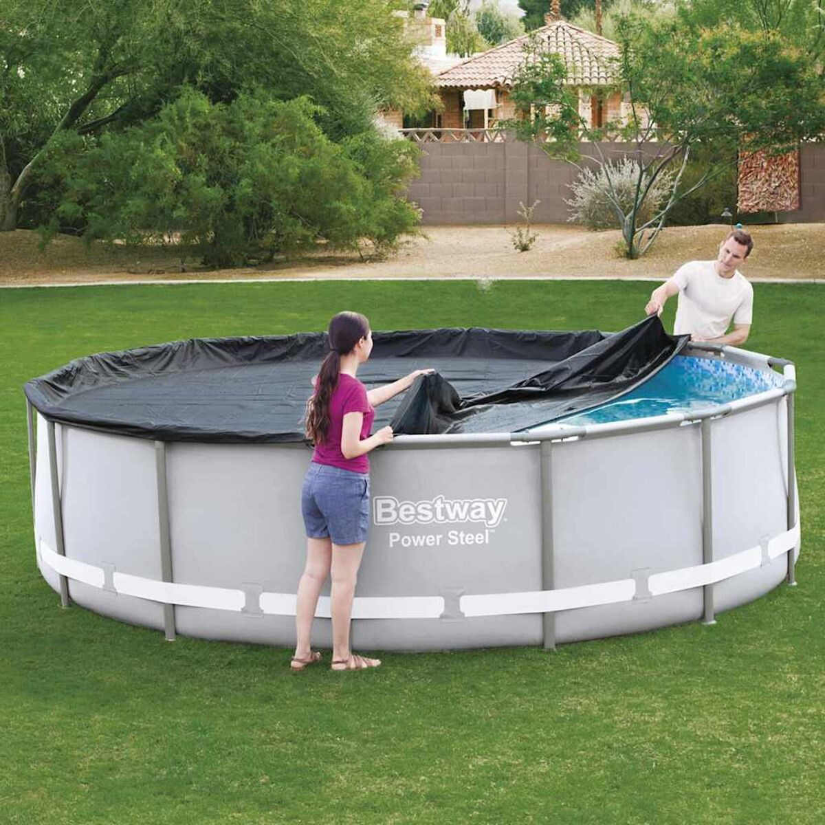 Personen bedecken einen Swimmingpool mit einer schwarzen Abdeckung. Grünes Gras und Häuser im Hintergrund.
