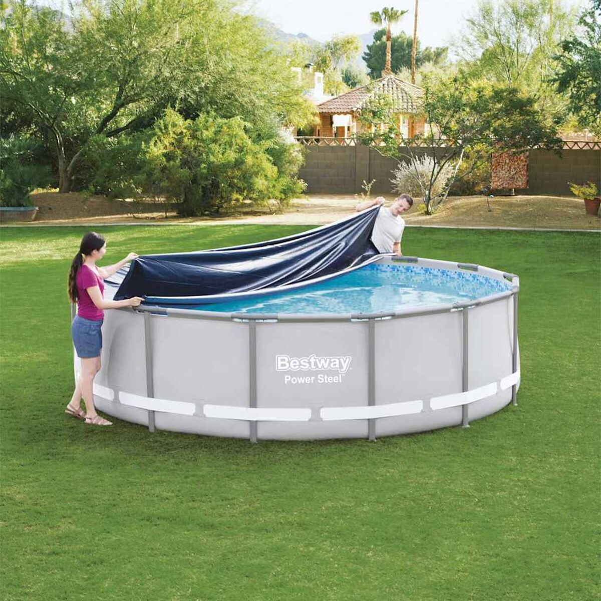 Zwei Personen bedecken einen blauen oberirdischen Pool mit einer schwarzen Abdeckung auf einem grünen Rasen.