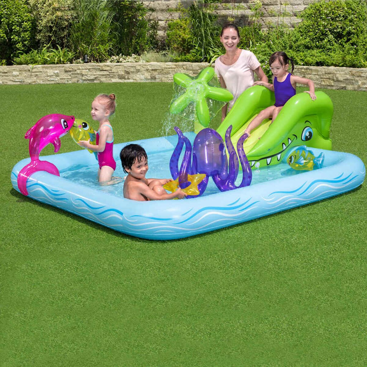 Kinder im Pool mit aufblasbaren Spielzeugen und Rutsche. Eine Frau beaufsichtigt sie am Rand.