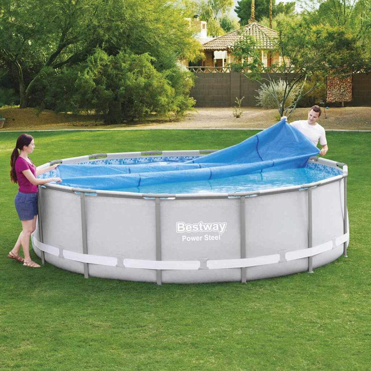 Zwei Personen decken einen runden Pool mit einer blauen Abdeckung ab. Grünes Gras umgibt den Pool, Bäume und ein Haus.