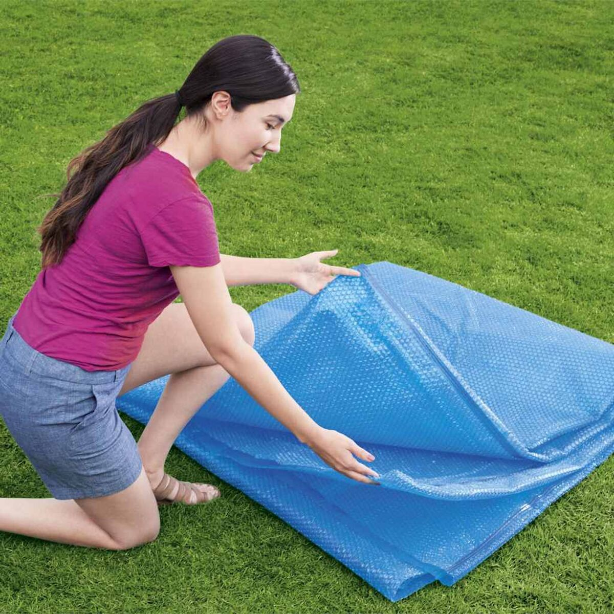 Eine Frau kniet auf dem Rasen und entfaltet eine blaue Poolabdeckung mit Luftpolsterfolie.
