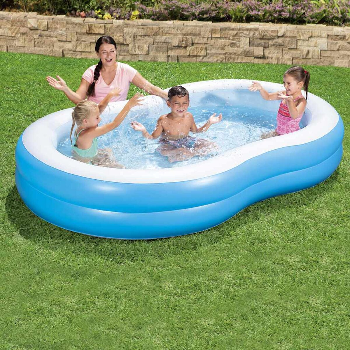 Vier Kinder spielen in einem blau-weißen aufblasbaren Pool auf einer Rasenfläche, im Hintergrund eine Frau.