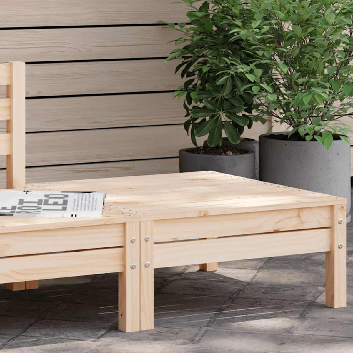 Lettino in legno per esterni. Basso, con una rivista e due piante in vaso sullo sfondo.