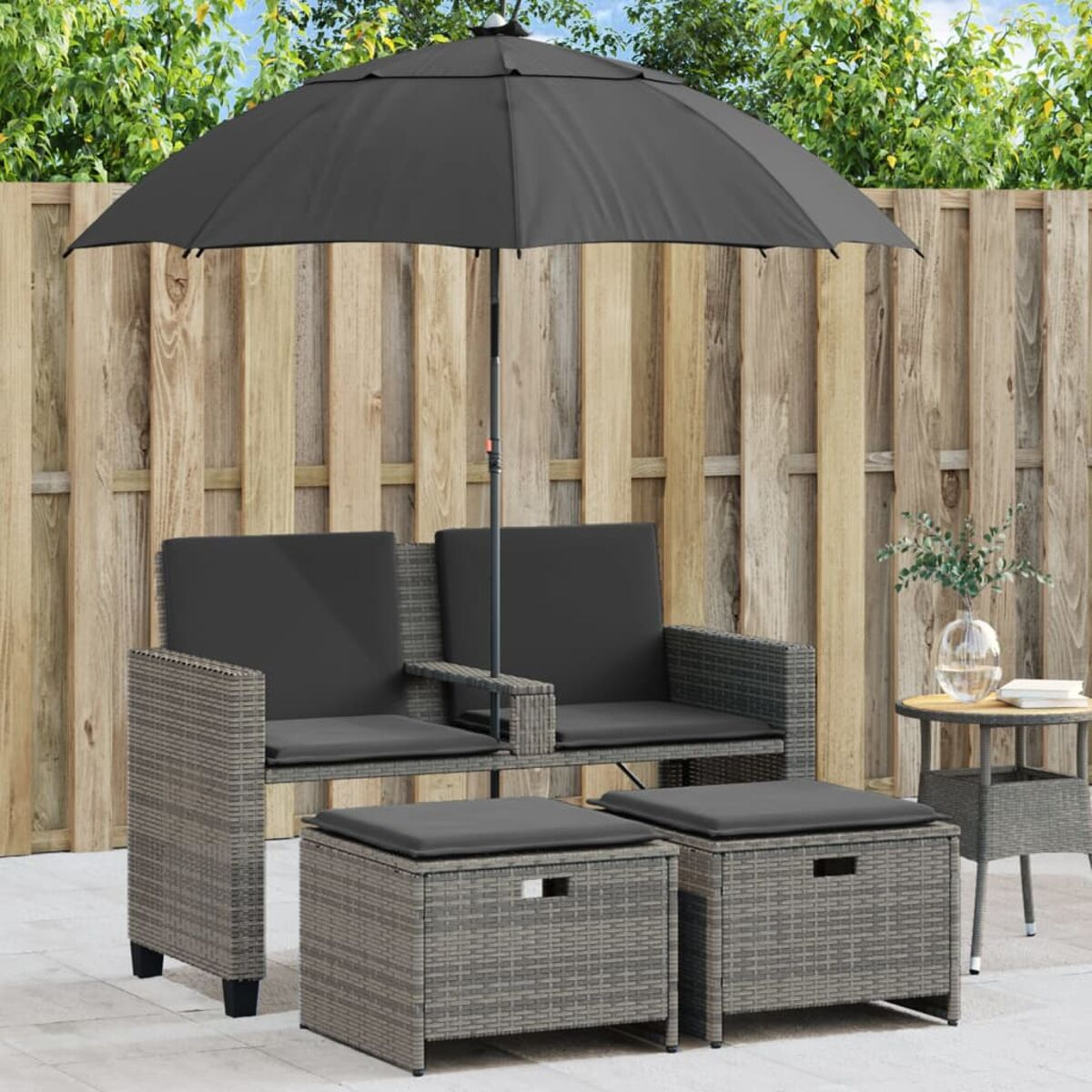 Un set di mobili con un divano in vimini grigio, due pouf e un ombrello nero. Su un patio.