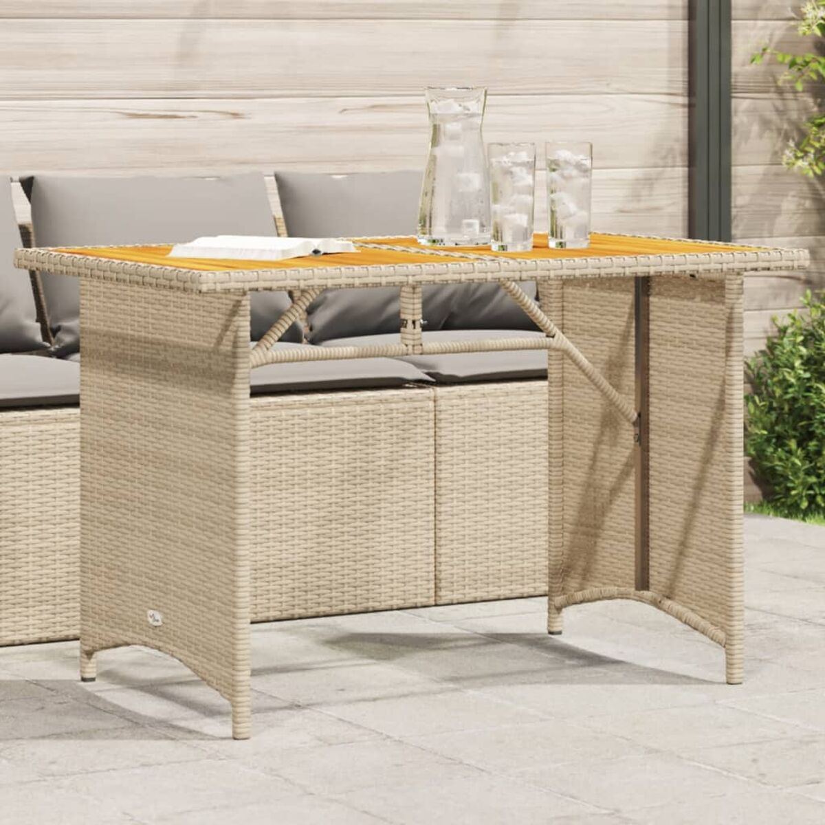 Wicker-Tisch mit Holzplatte, Wasserkrug und Gläsern im Freien. Hintergrund: Korbsessel und Holzwand.