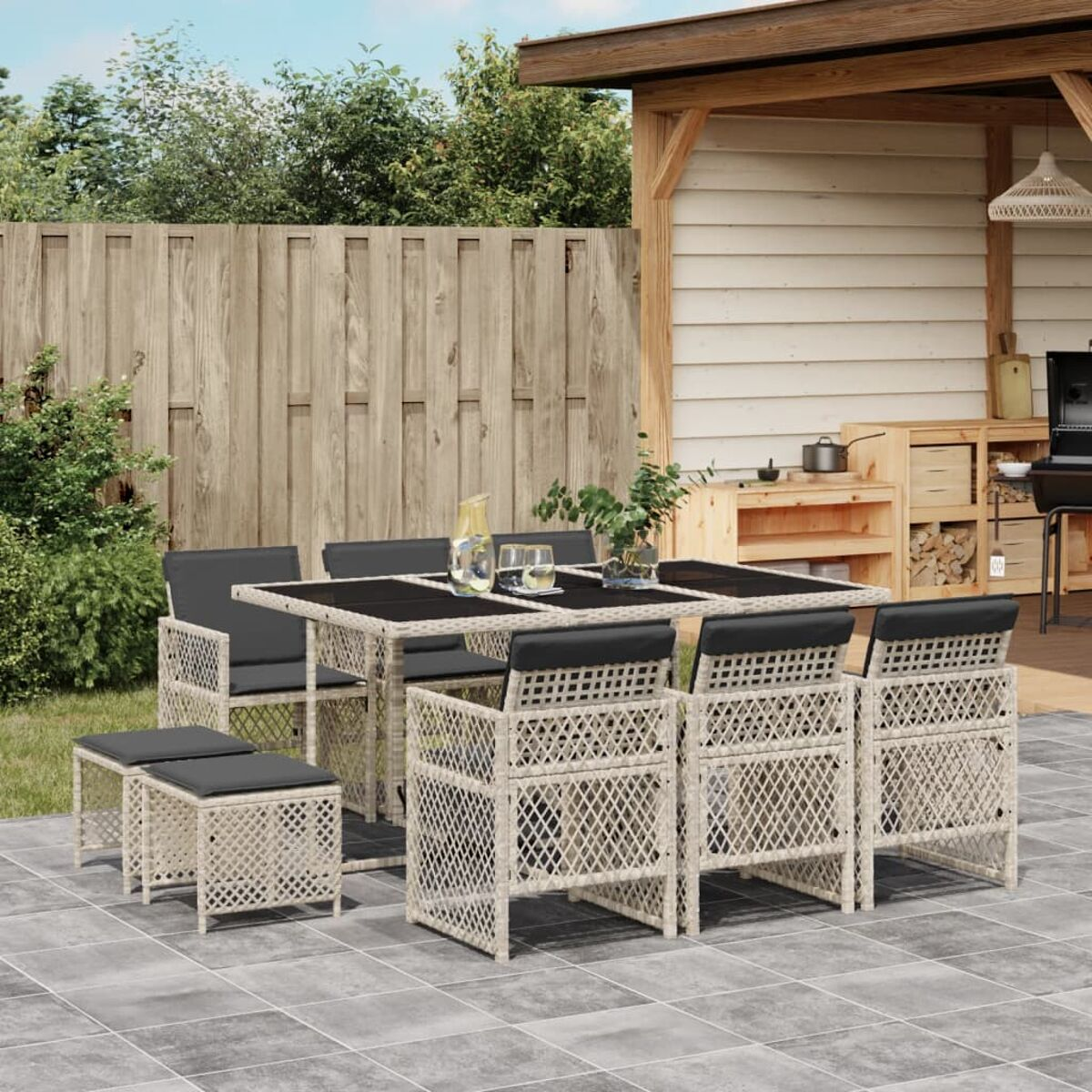 Set patio: tavolo, sedie e poggiapiedi. Vimini beige con cuscini neri. Recinzione in legno, capannone dietro.