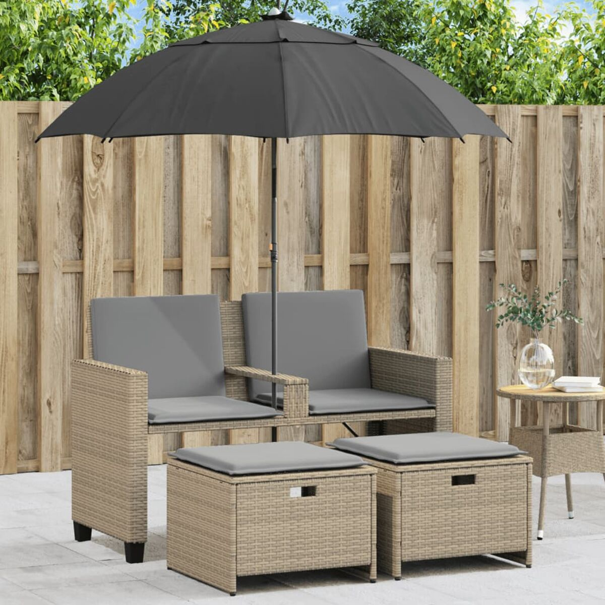 Set di sedute per esterni. Mobili in rattan beige, cuscini grigi, ombrellone e recinzione in legno.
