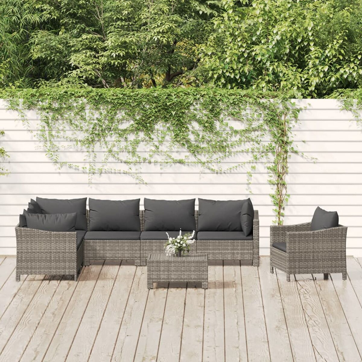 Graues Rattan-Terrassenset mit Kissen, einem Tisch und einem weißen Zaun mit Grün.