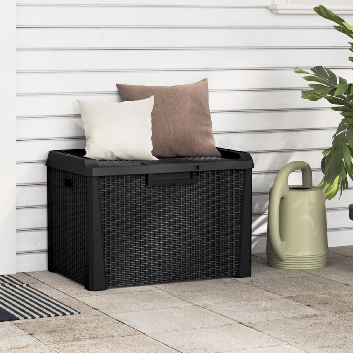 Schwarze Outdoor-Aufbewahrungsbox mit Webmuster. Zwei Kissen oben. Eine Gießkanne ist an der Seite.