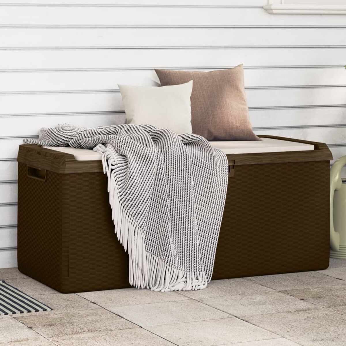 Braune Aufbewahrungsbox für den Außenbereich mit weißer Decke und Kissen. Eine Gießkanne befindet sich im Hintergrund.