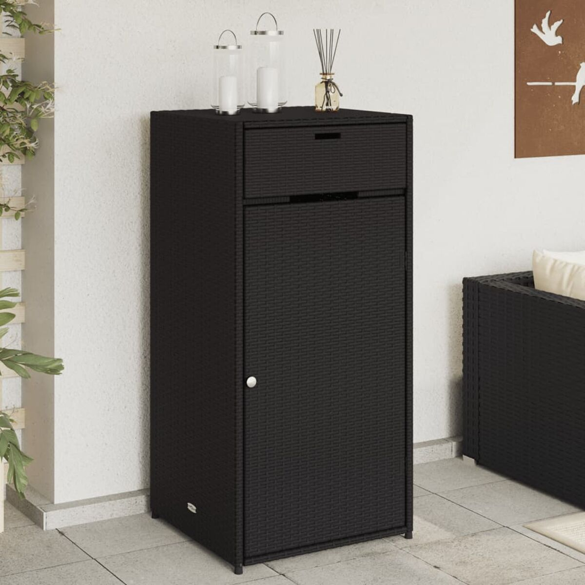 Ein schwarzer Schrank aus Korbgeflecht mit Schublade und Tür. Er steht auf einer Terrasse.