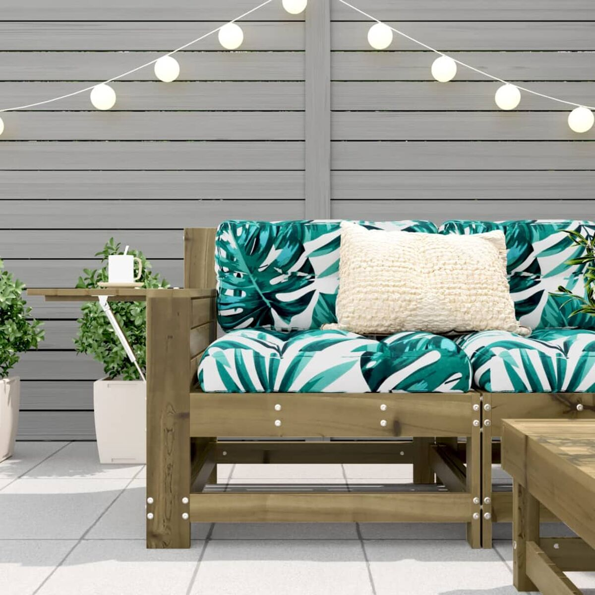 Outdoor-Sofa mit tropischen Blattkissen und einem kleinen Beistelltisch auf einer Terrasse mit Lichterketten.