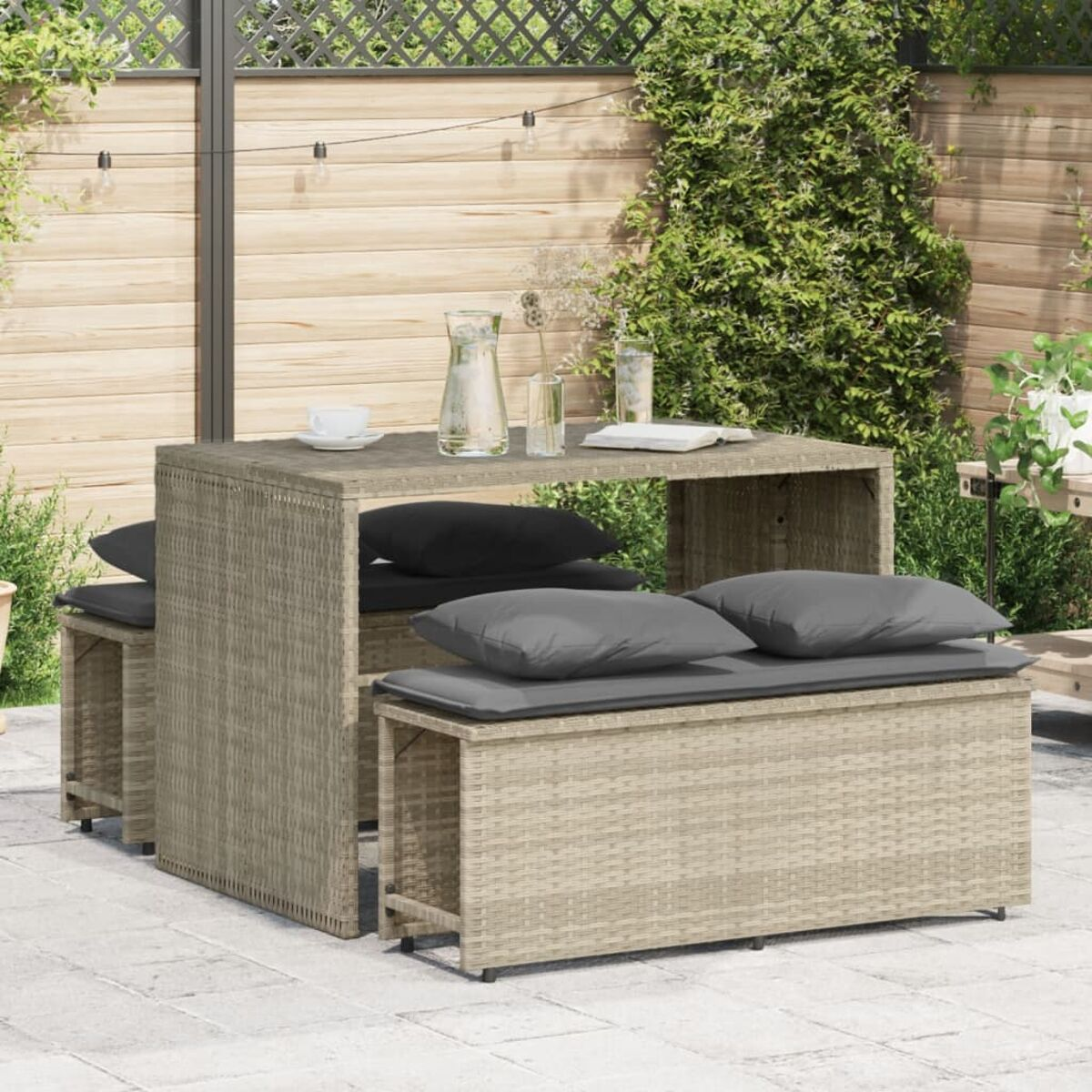 Wicker-Tisch und -Bänke. Graue Kissen auf den Bänken. Pflanzen und ein Zaun im Hintergrund.