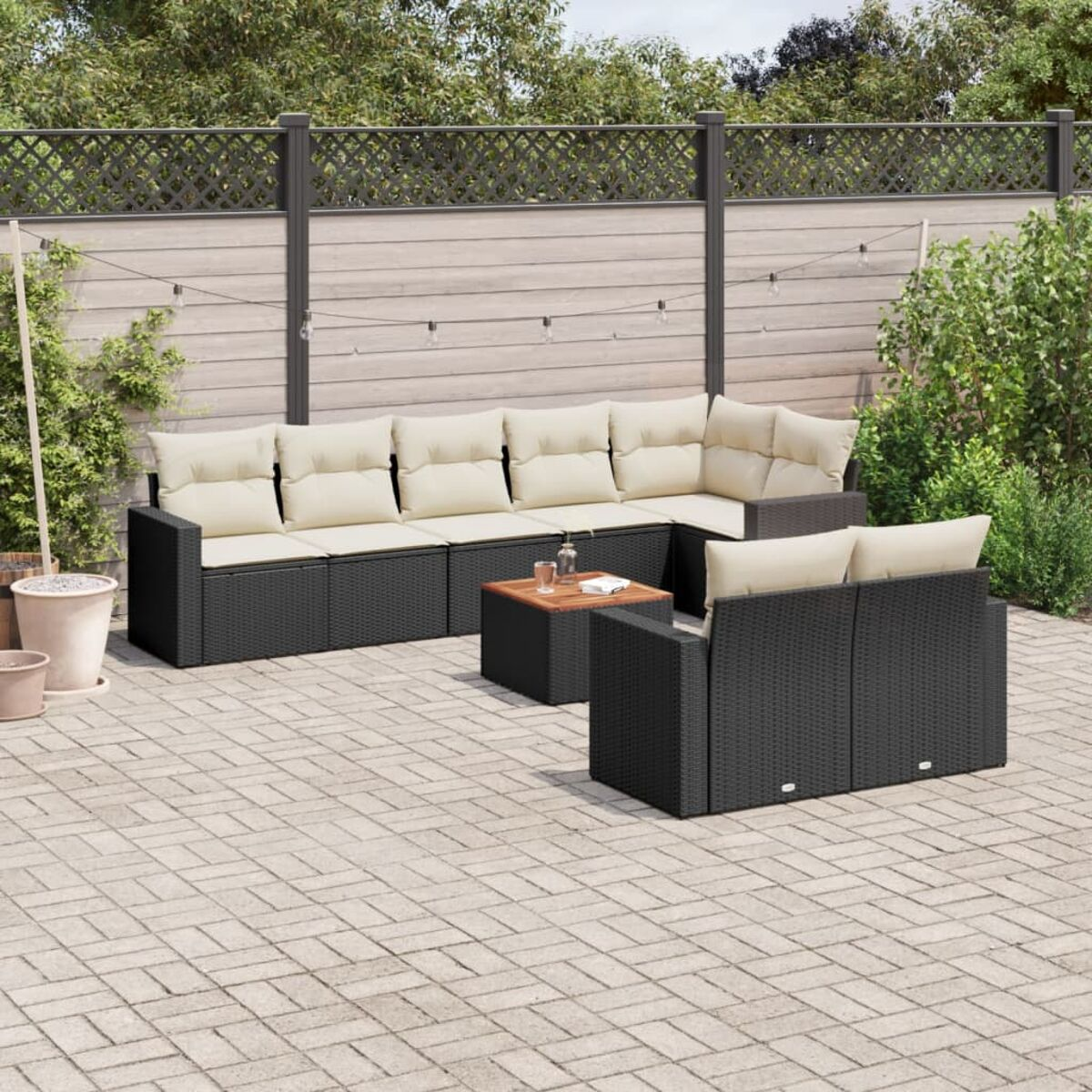 Schwarzes Rattan-Ecksofa mit beigen Kissen. Holzzaun und Grün im Hintergrund.