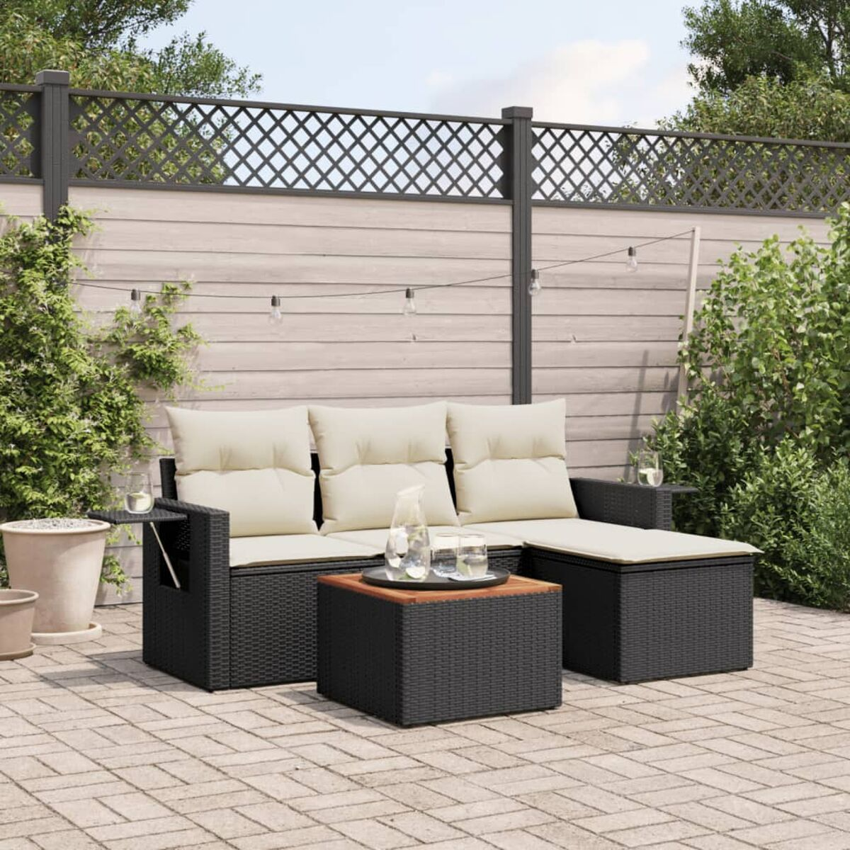 Schwarzes Ecksofa aus Korbgeflecht für draußen mit cremefarbenen Kissen, Holztisch und Ziegelterrasse. Garten im Hintergrund.