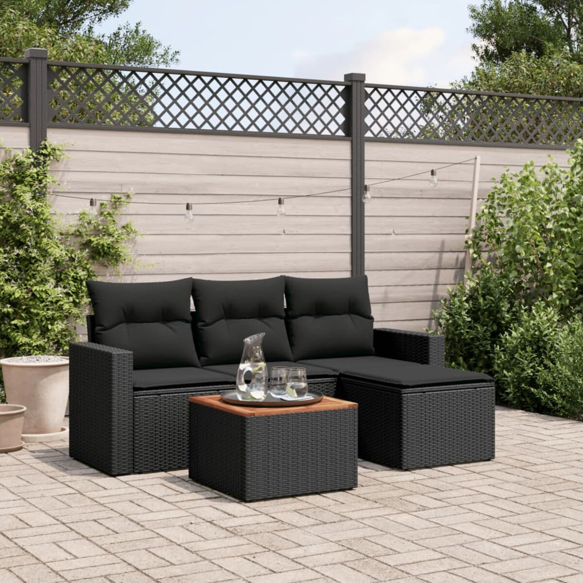 Schwarzes Outdoor-Ecksofa und Tisch-Set aus Korbgeflecht auf einer Terrasse mit Pflanzen und Lichterketten.