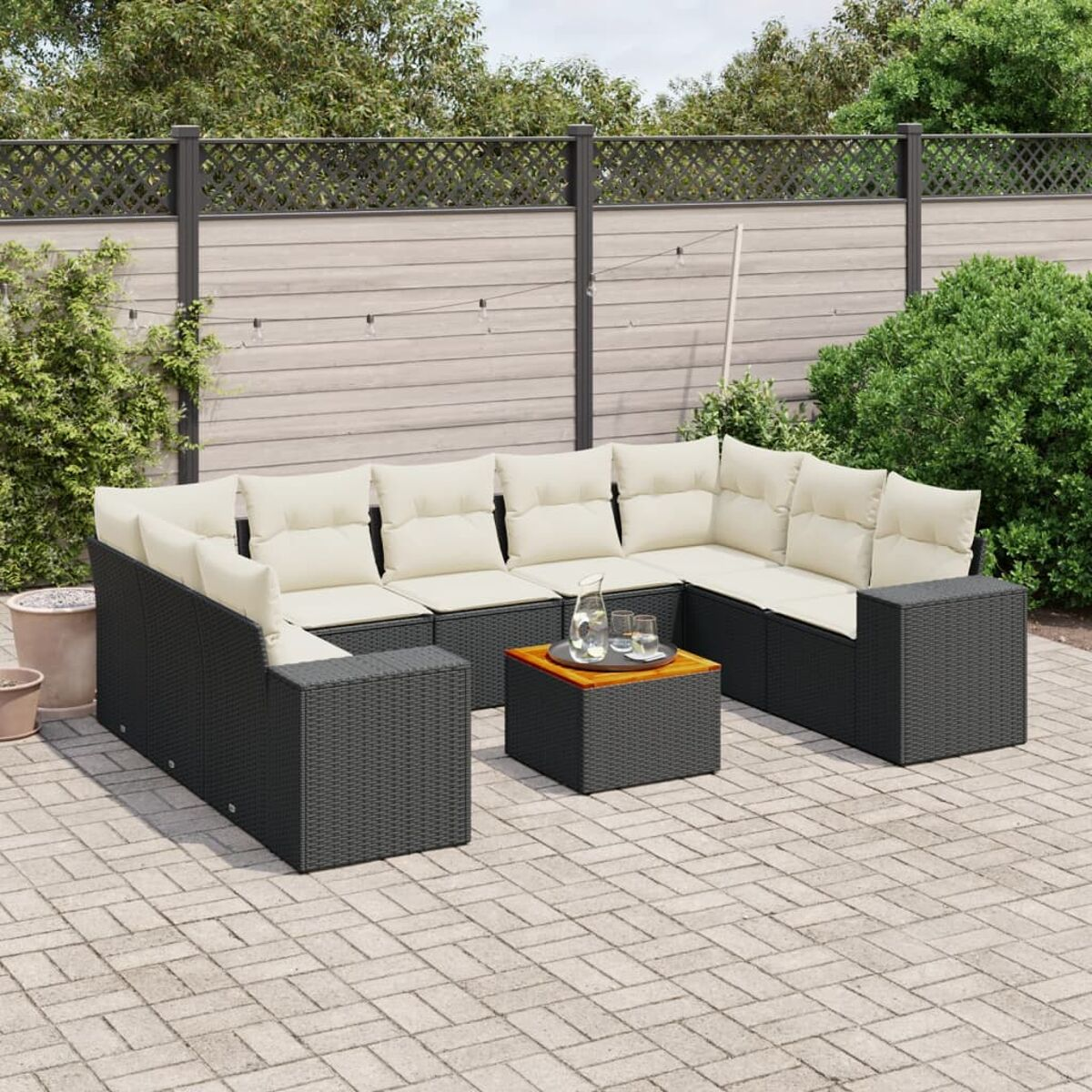 Schwarzes Outdoor-Rattan-Sofaset mit beigen Kissen, Tisch. Ziegelterrasse, Zaun, Pflanzen.