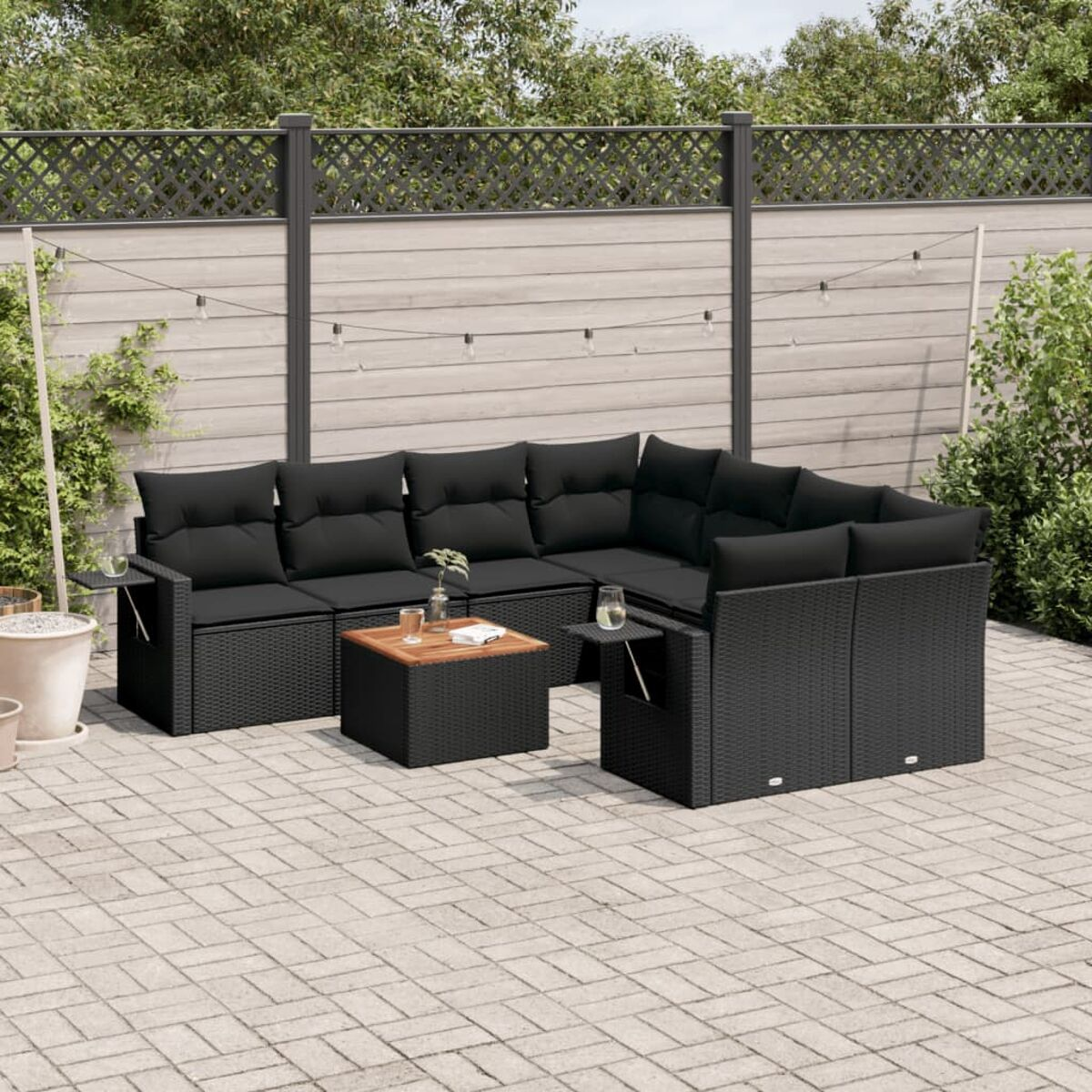 Schwarzes Ecksofa im Freien mit schwarzen Kissen. Ein quadratischer Tisch davor. Gezäunter Hintergrund mit Pflanzen.