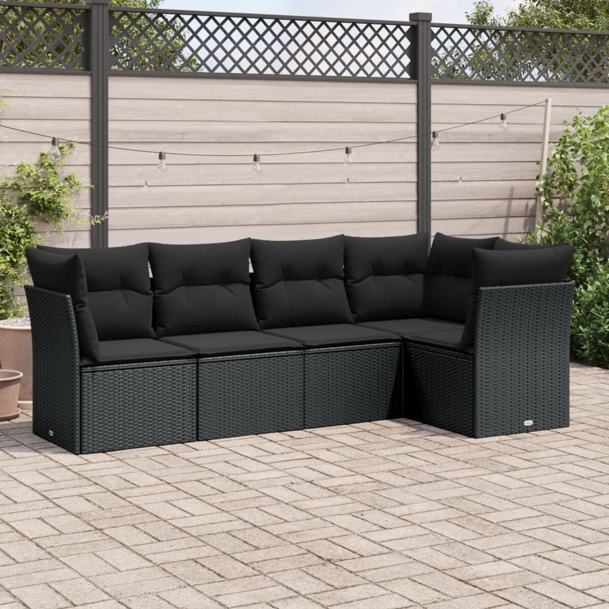 Schwarzes Ecksofa aus Rattan mit schwarzen Kissen auf einer Backstein-Terrasse, vor einem Holzzaun mit Lichtern.