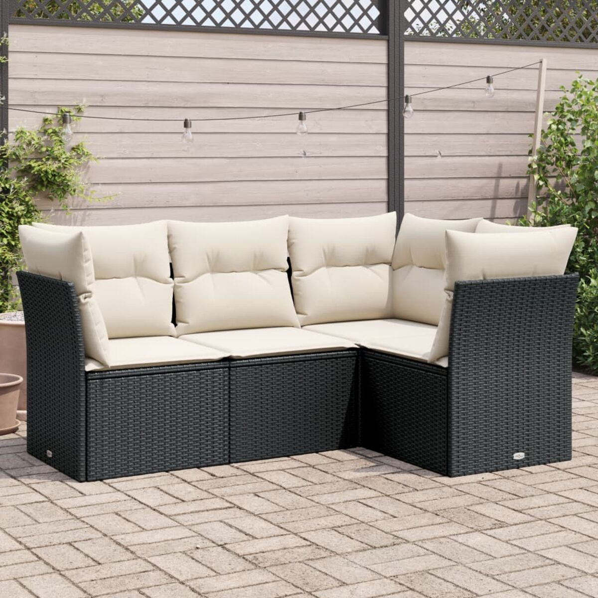 Schwarze Wicker-Sofagarnitur mit weißen Kissen. Ziegelterrasse, Holzzaun und Lichterkette im Hintergrund.