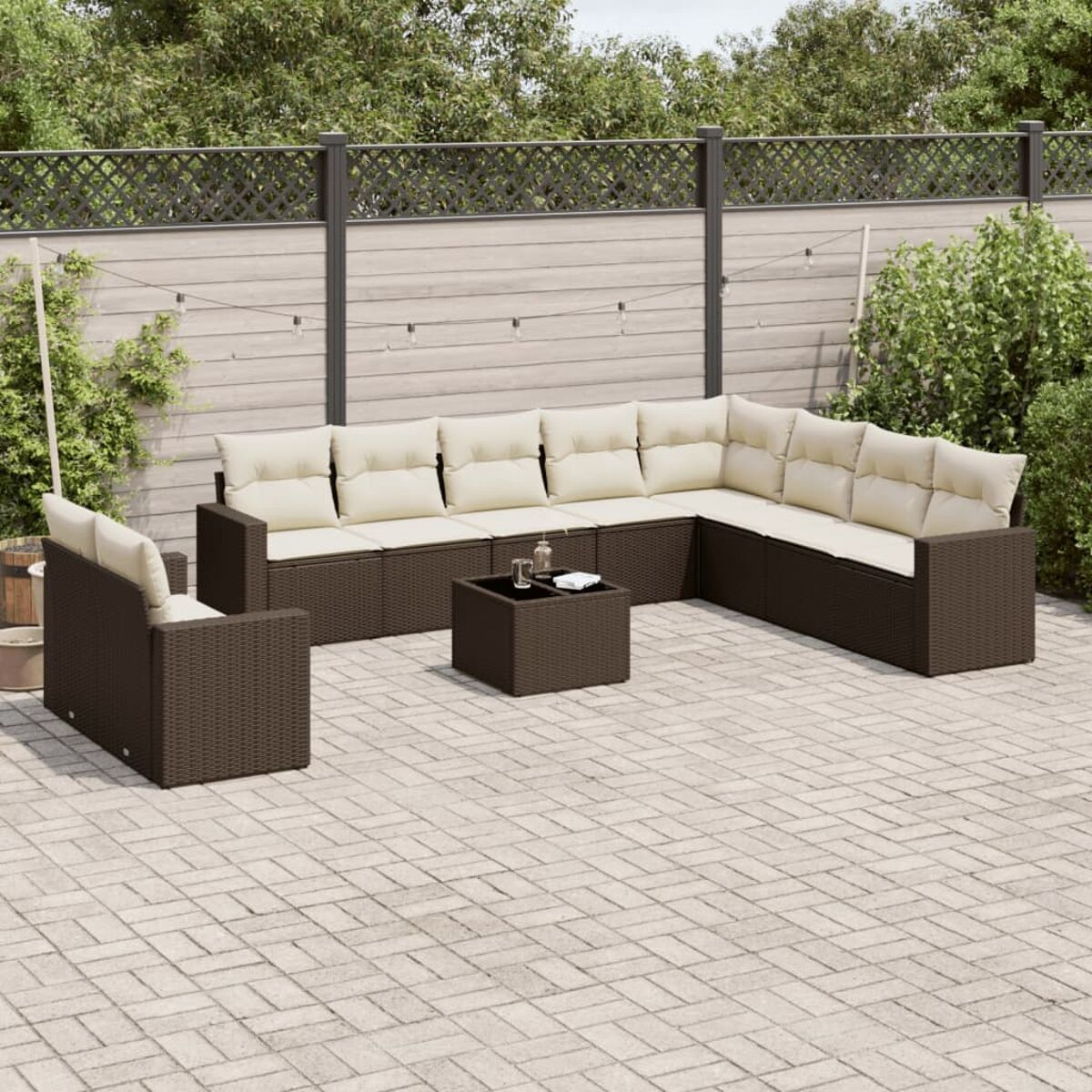 Divano angolare marrone da esterno con cuscini beige. Tavolo quadrato davanti. Patio in mattoni con recinzione e piante.
