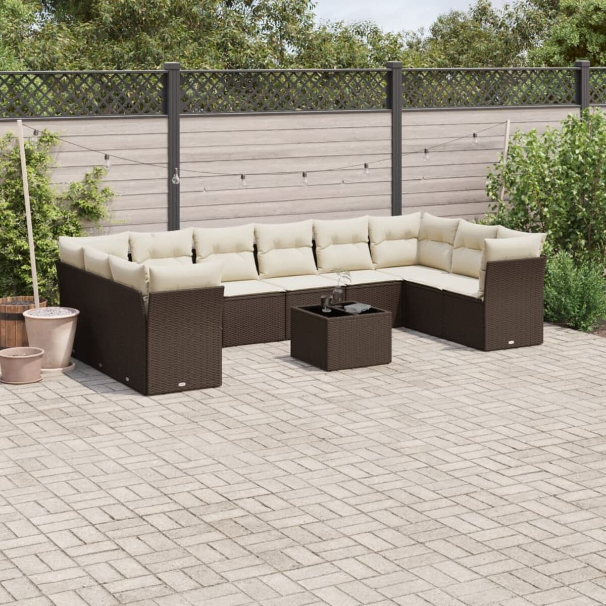 Set divano in vimini marrone con cuscini color crema su un patio. Recinzione in legno, piante e luci sullo sfondo.