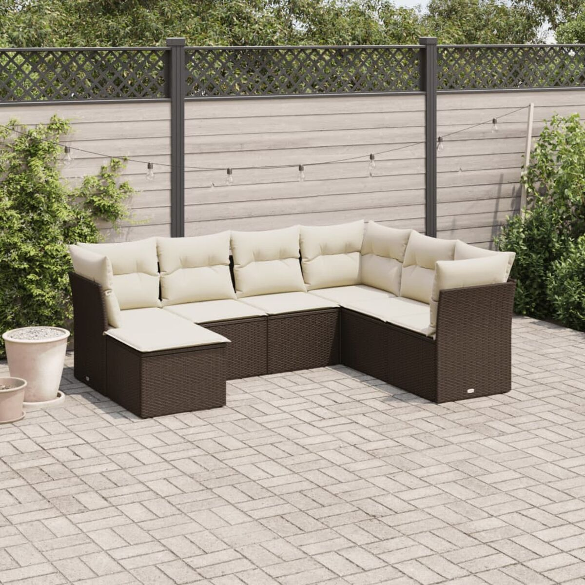 Braunes Geflecht-Sofaset mit cremefarbenen Kissen, auf einer Terrasse mit Zaun und Büschen. Lichterketten oben.