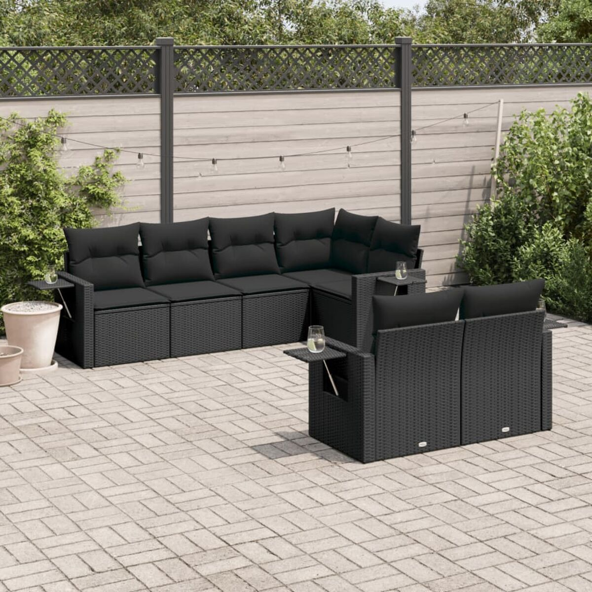 Schwarzes Ecksofa im Freien. Ein quadratischer Tisch davor. Umzäunter Hintergrund mit Pflanzen. Backstein-Terrasse.