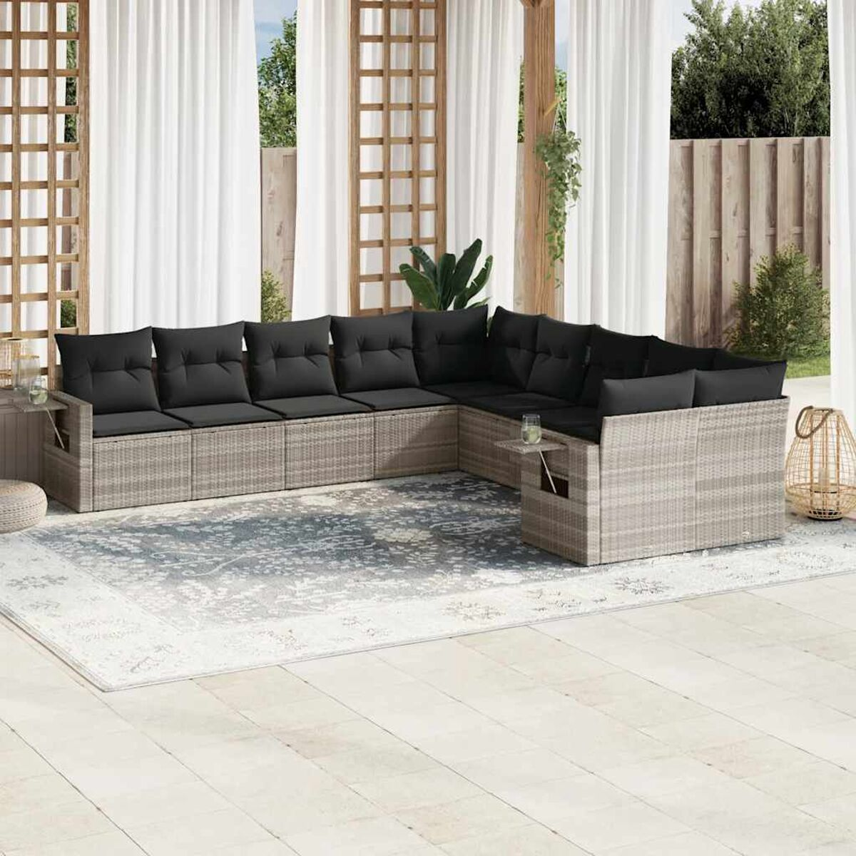 Ein Outdoor-Ecksofa aus Rattan mit schwarzen Kissen. Das Set befindet sich auf einer Terrasse mit Teppich und Vorhängen.