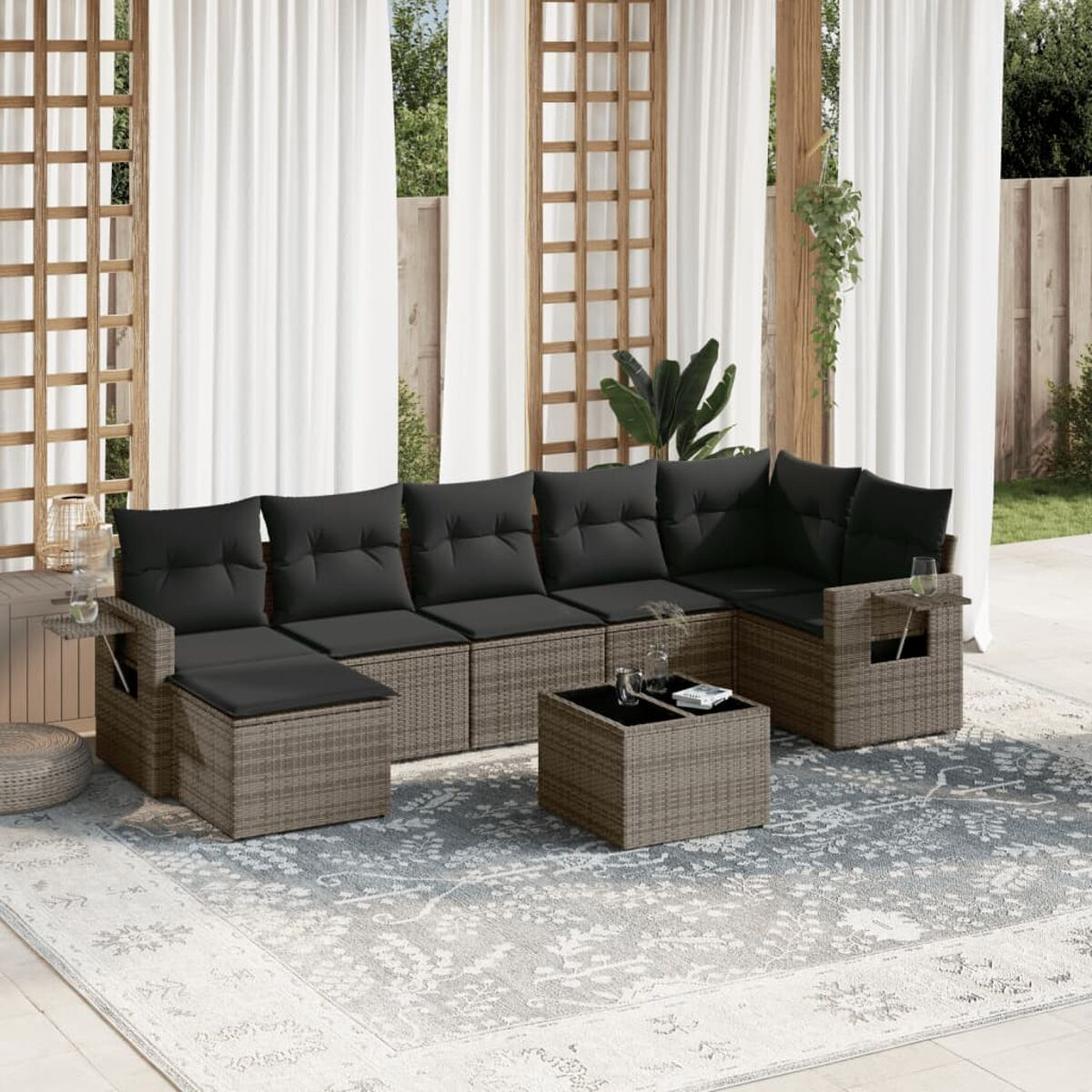 Terrassenset mit grauen Korbsesseln, schwarzen Kissen, Tisch und gemustertem Teppich. Weiße Vorhänge.