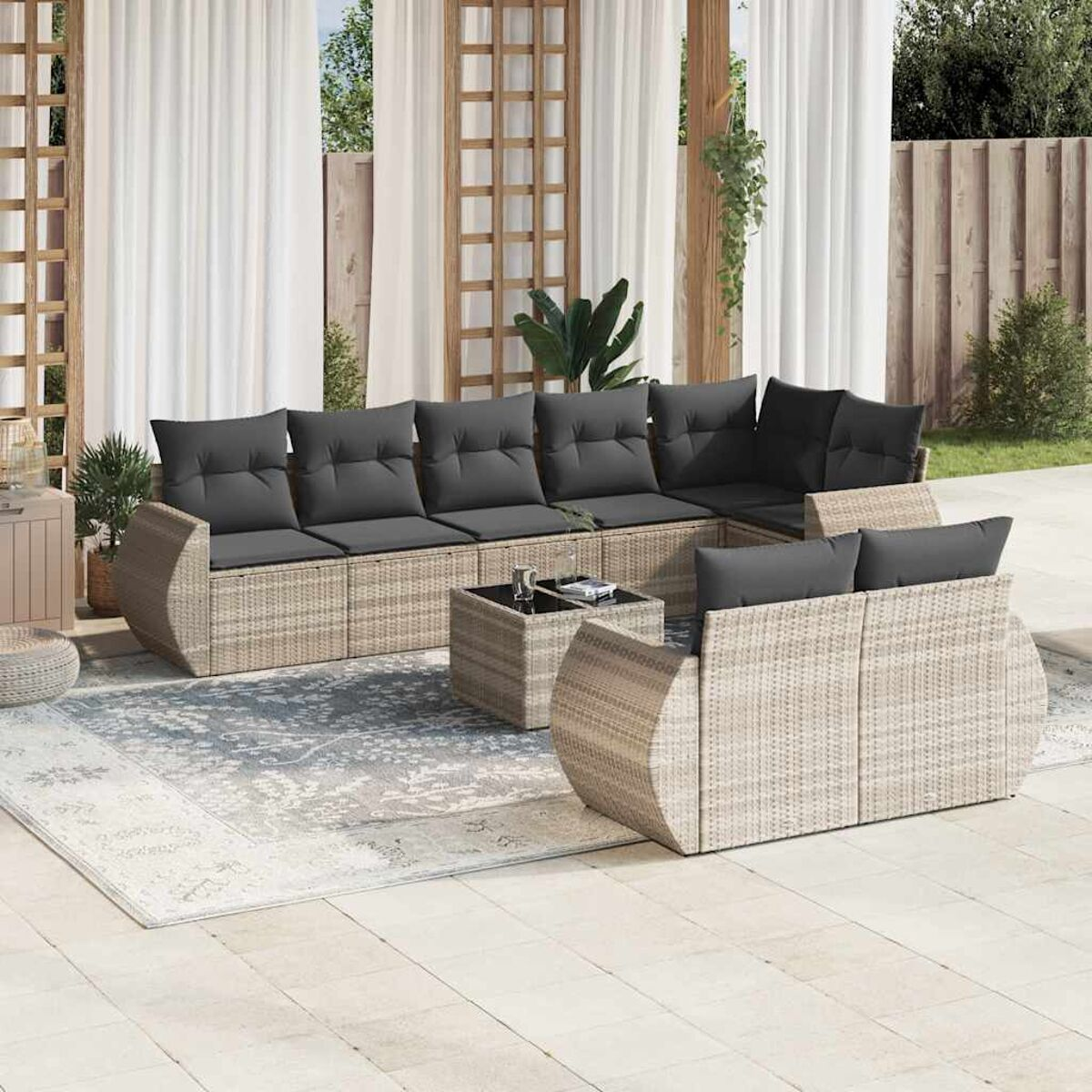 Ein helles Wicker-Ecksofa mit dunkelgrauen Kissen auf einer Terrasse mit Teppich und Pflanzen.