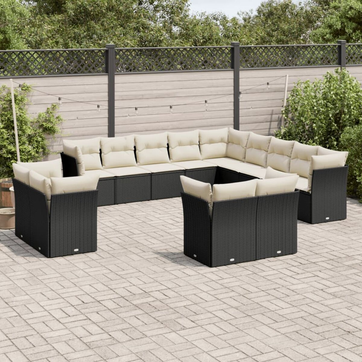 Schwarzes Ecksofa im Freien. Ein quadratischer Tisch davor. Backstein-Terrasse mit Zaun und Pflanzen.