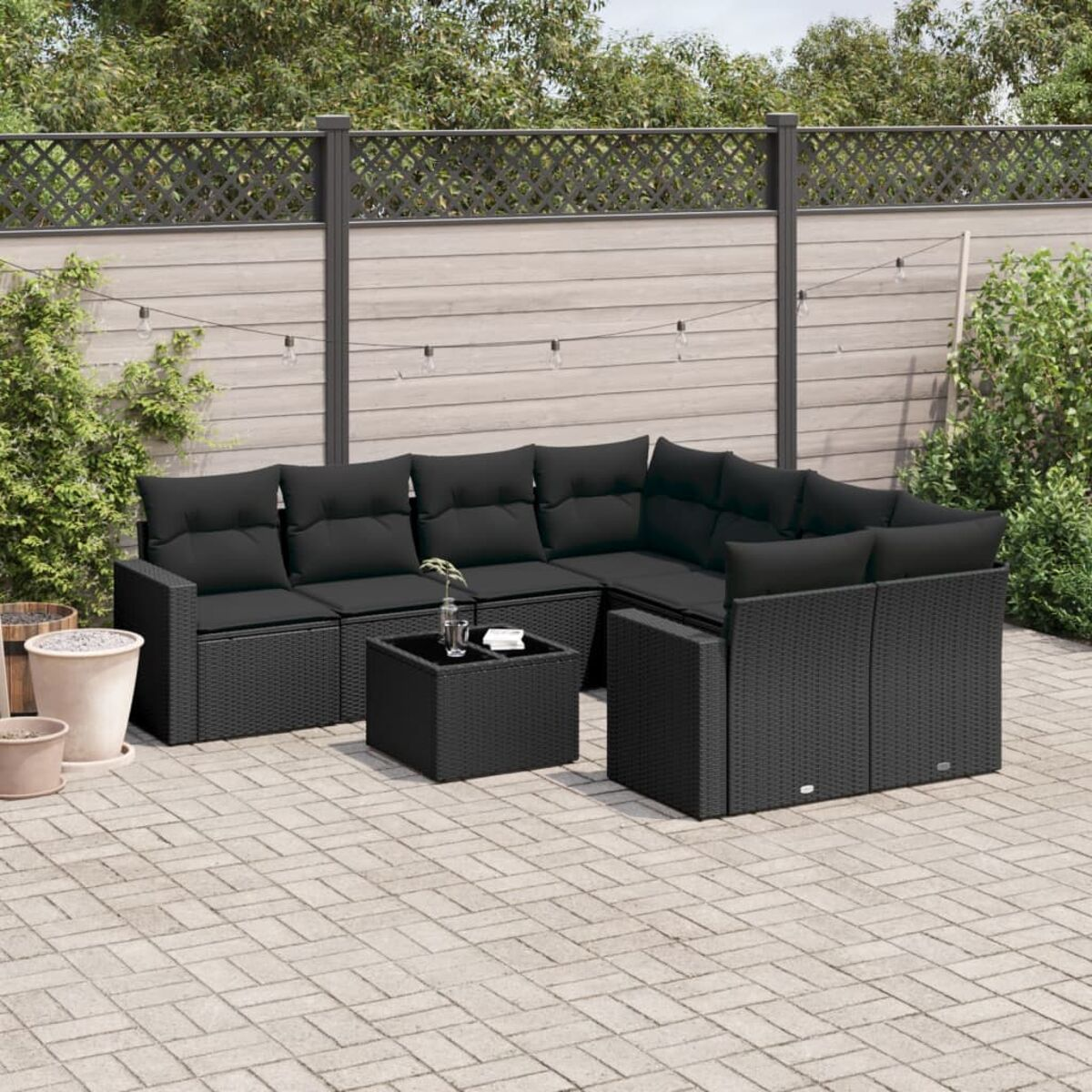 Schwarzes Outdoor-Ecksofa aus Korbgeflecht mit schwarzen Kissen, auf einer Backstein-Terrasse, mit Grün und Zaun.