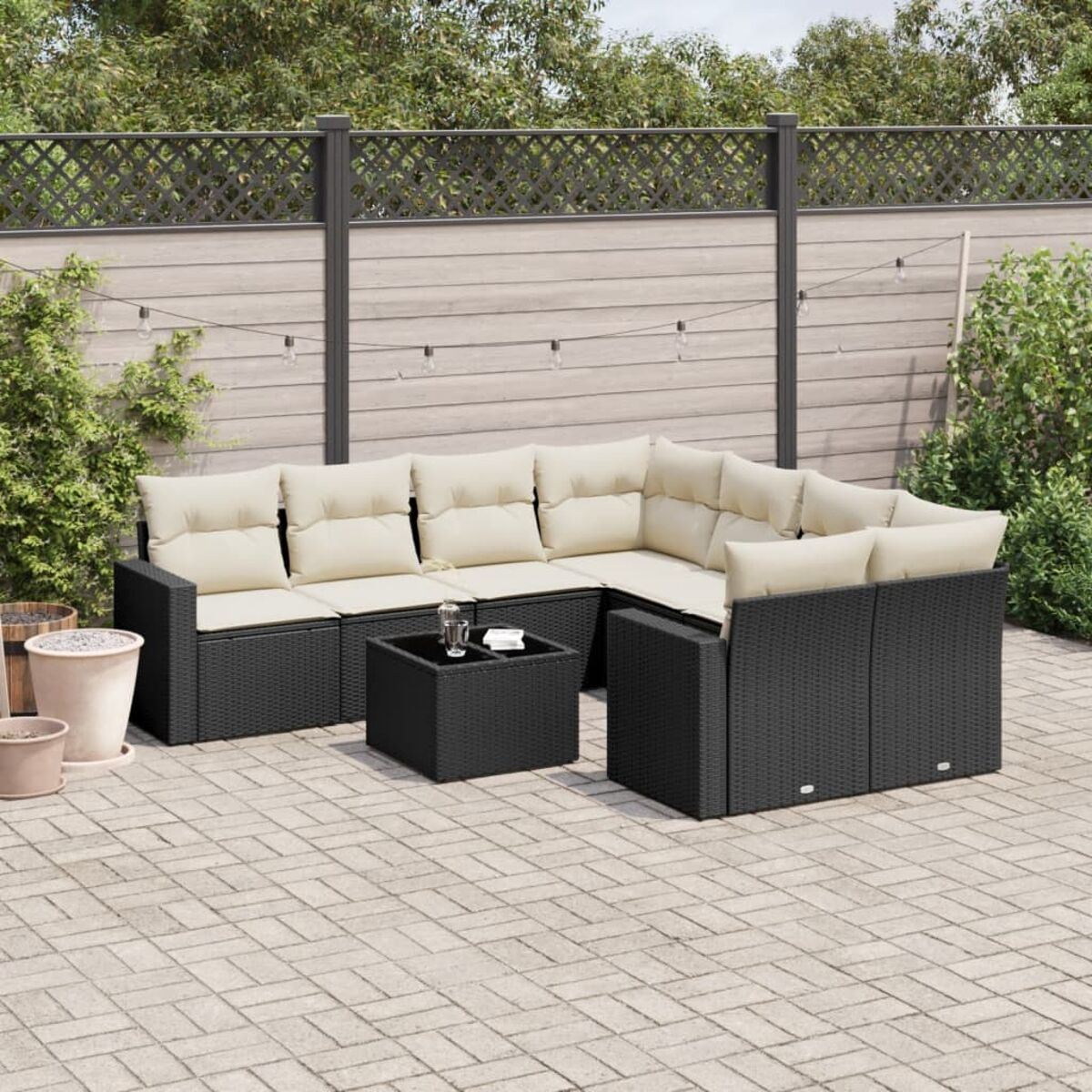 Schwarzes Outdoor-Ecksofa aus Korbgeflecht mit beigefarbenen Kissen, einem passenden Tisch, auf einer Ziegelterrasse und einem Zaun im Hintergrund.