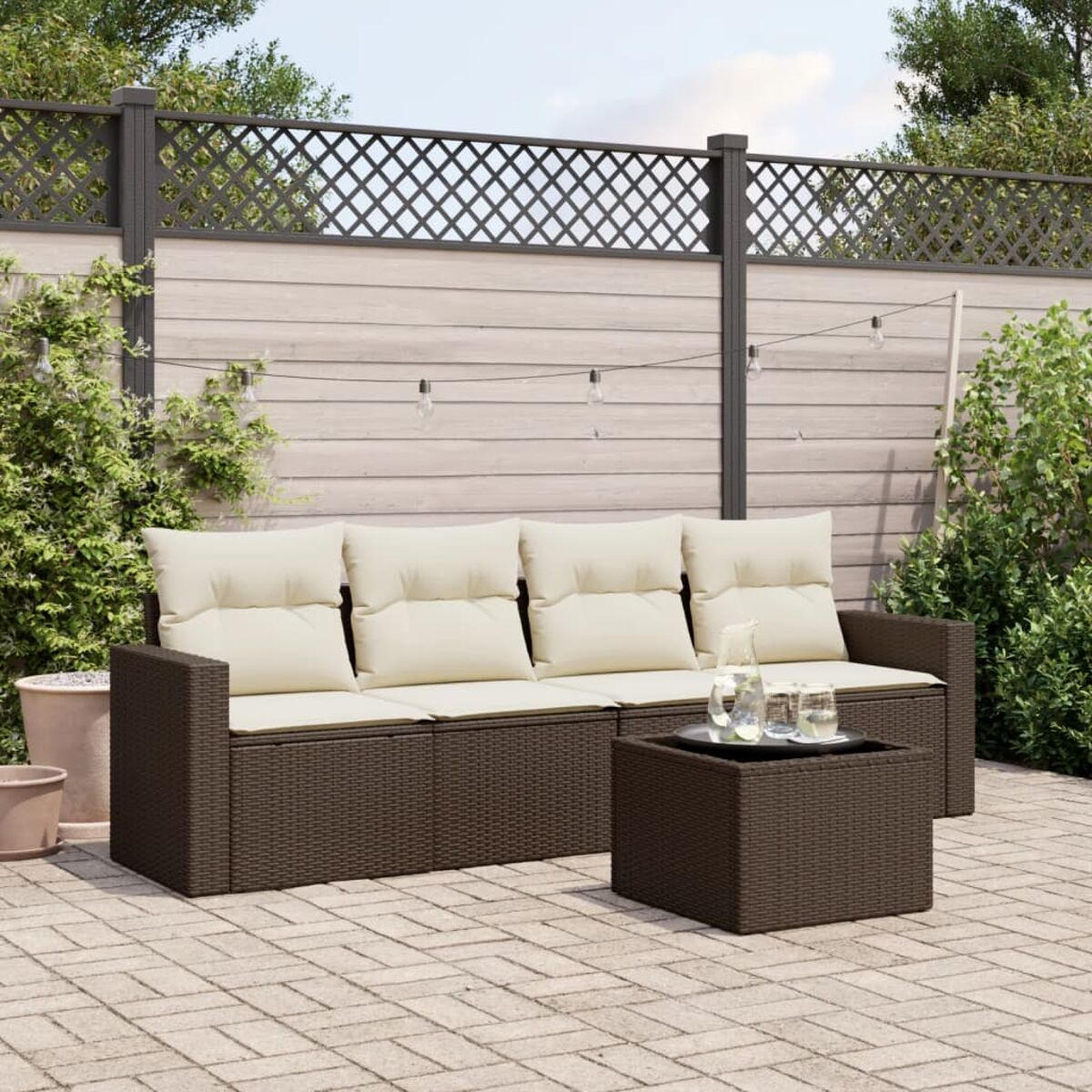 Set divano in vimini marrone da esterno con cuscini beige, un tavolino e una recinzione a traliccio sullo sfondo.