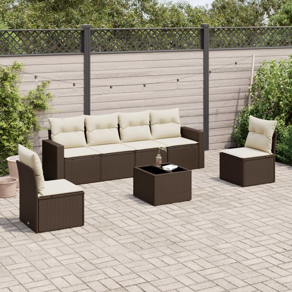 Set di sedute da esterno in vimini marrone con cuscini beige, un tavolo, su un patio in mattoni, con recinzione e vegetazione.