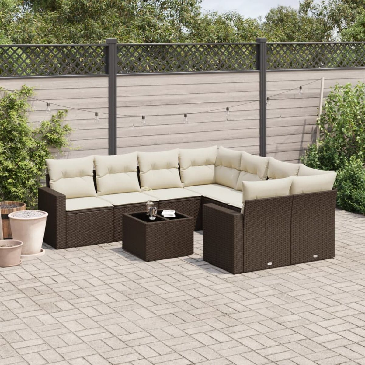 Divano in rattan marrone con cuscini color crema. Il set è su un patio in mattoni. Piante e una recinzione sullo sfondo.