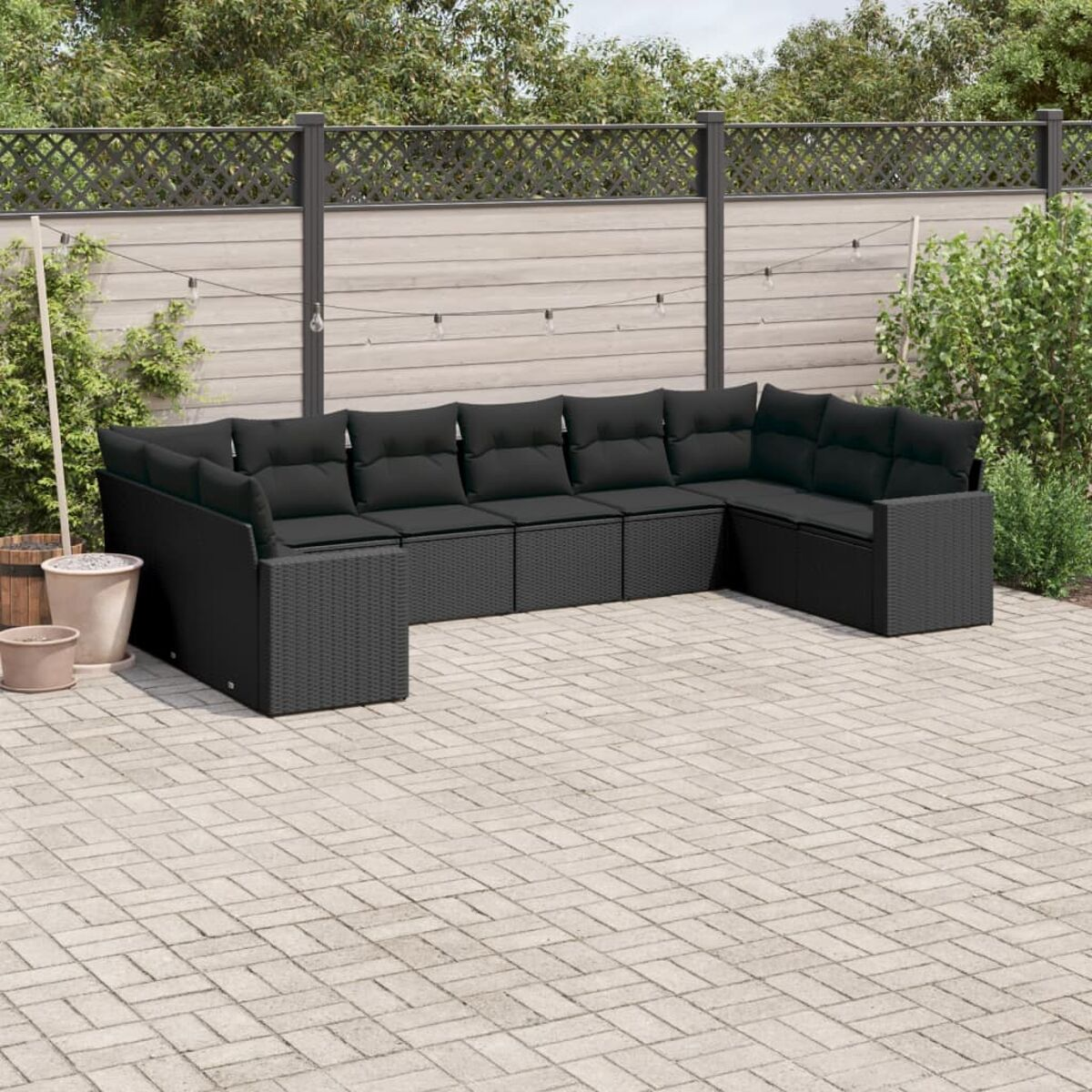 Schwarzes Rattan-Ecksofa mit schwarzen Kissen auf einer Backstein-Terrasse, mit Holzzaun im Hintergrund.