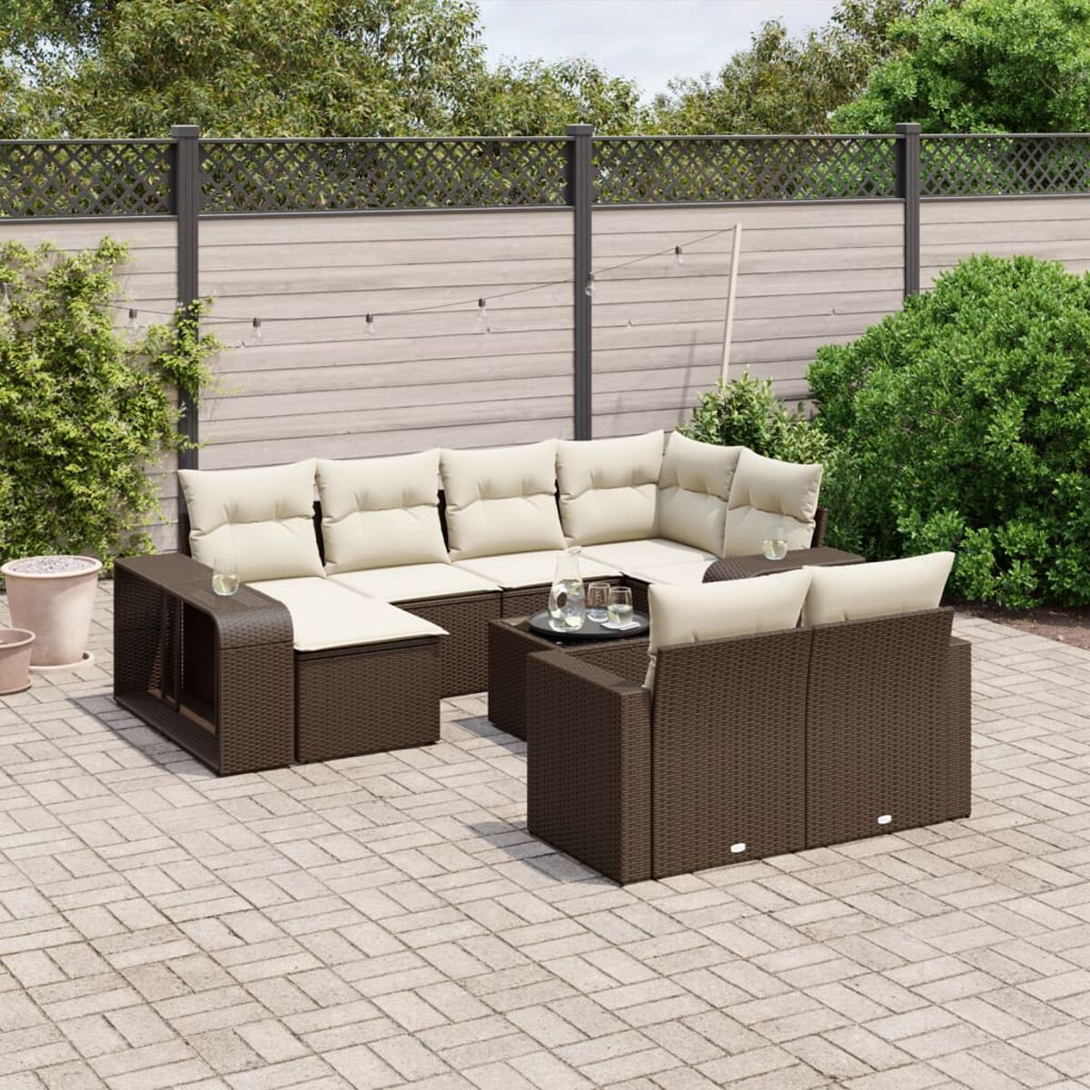 Set di mobili da patio in vimini marrone con cuscini beige, un tavolino e bevande. Patio in mattoni, cespugli verdi sullo sfondo.