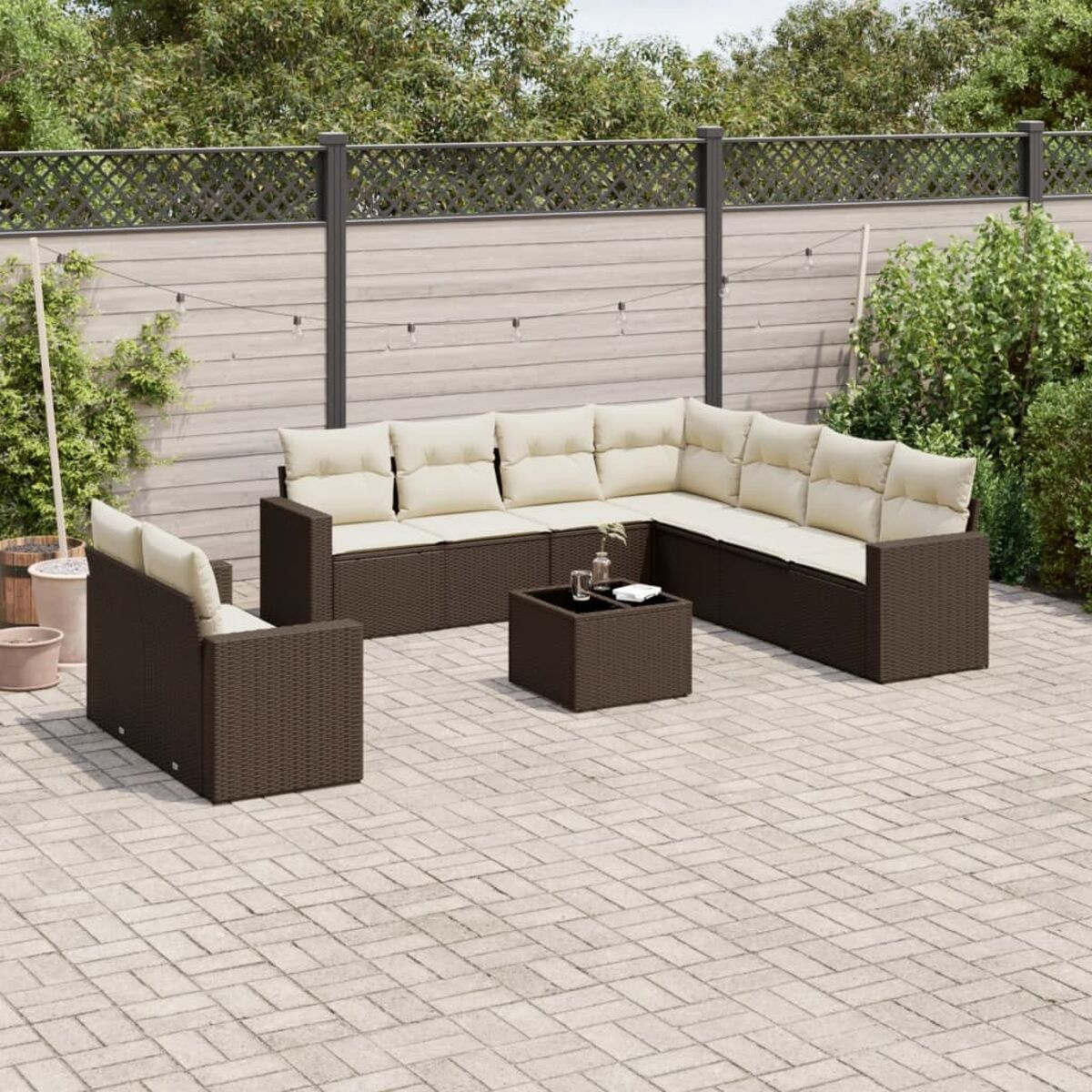 Set di mobili da patio in vimini marrone con cuscini color crema, su un patio in mattoni. Piante verdi e recinzione.