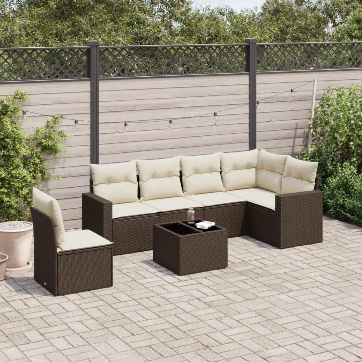 Set divano in rattan marrone con cuscini color crema e un tavolo. Il set è su un patio in mattoni con piante.