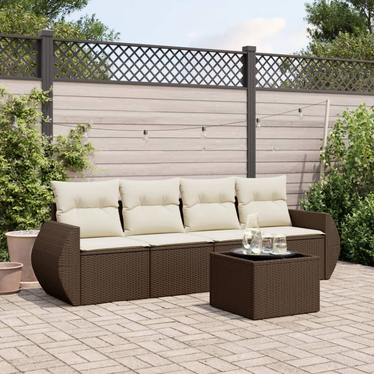 Set divano in vimini marrone con cuscini color crema e un tavolo nero. Patio pavimentato, sfondo recinzione.
