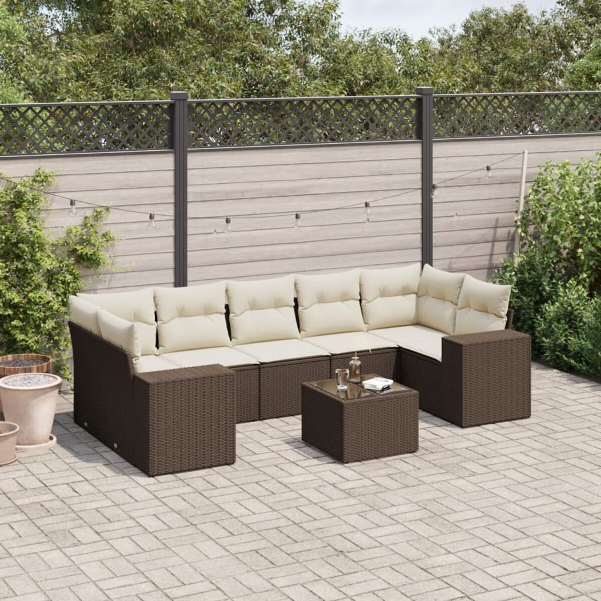 Set da patio in vimini marrone con cuscini color crema. Include divano, tavolino e piante circostanti.