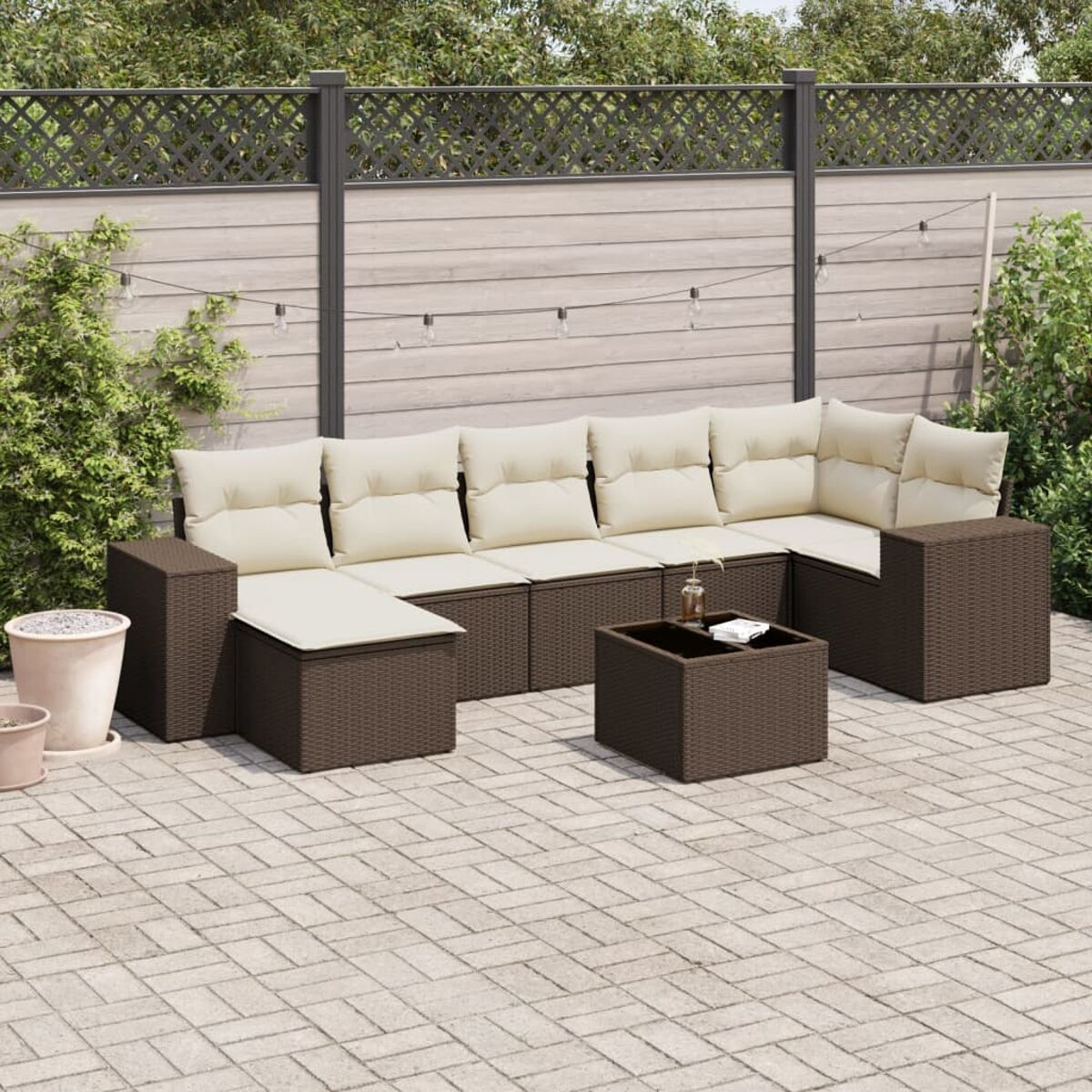 Divano in rattan marrone con cuscini color crema. Il set è su un patio in mattoni. Piante e una recinzione sullo sfondo.