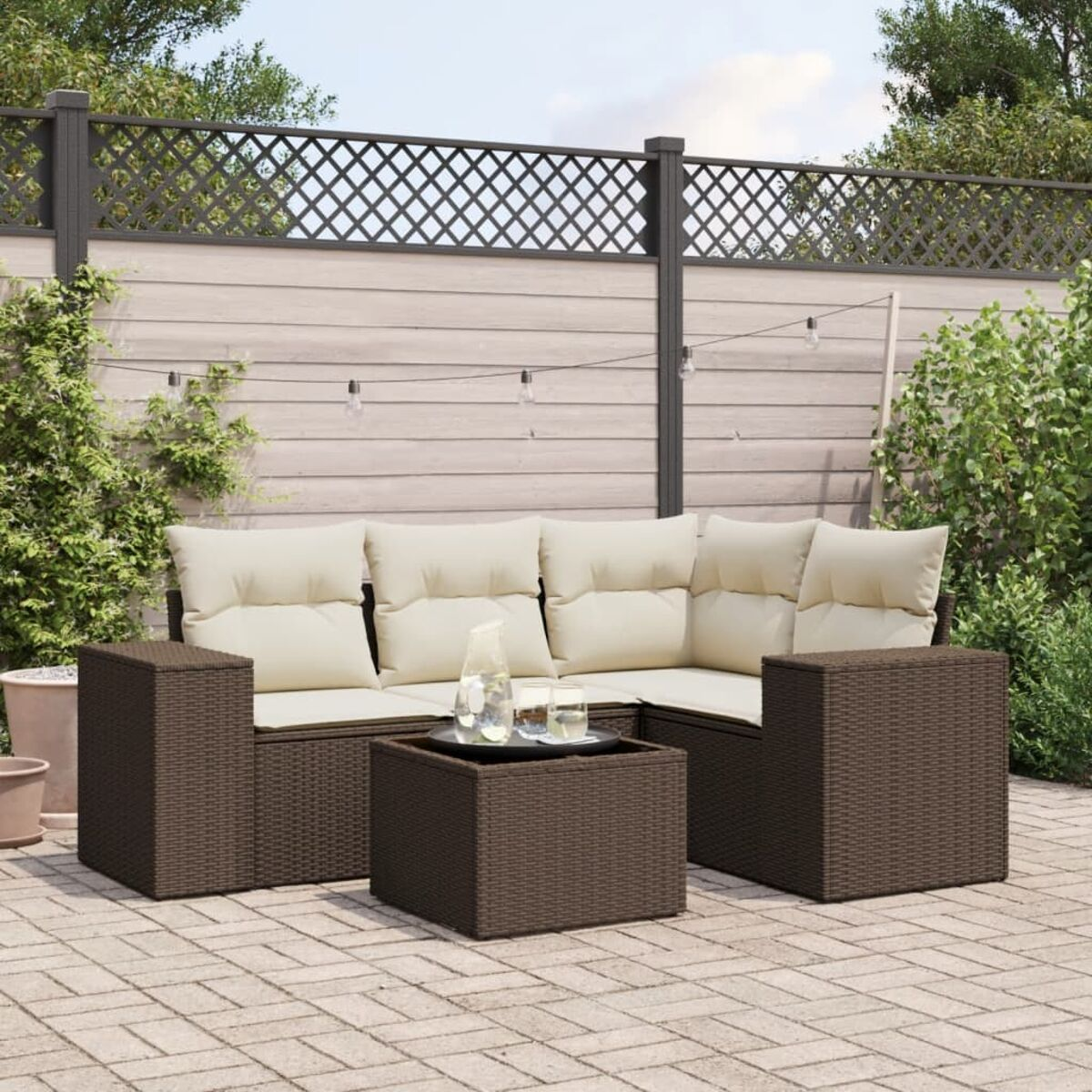 Divano angolare in vimini marrone con cuscini beige e tavolino su un patio in mattoni. Giardino sullo sfondo.
