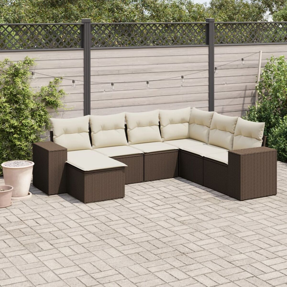 Divano angolare da esterno marrone con cuscini beige su un patio in mattoni, con recinzione e piante sullo sfondo.