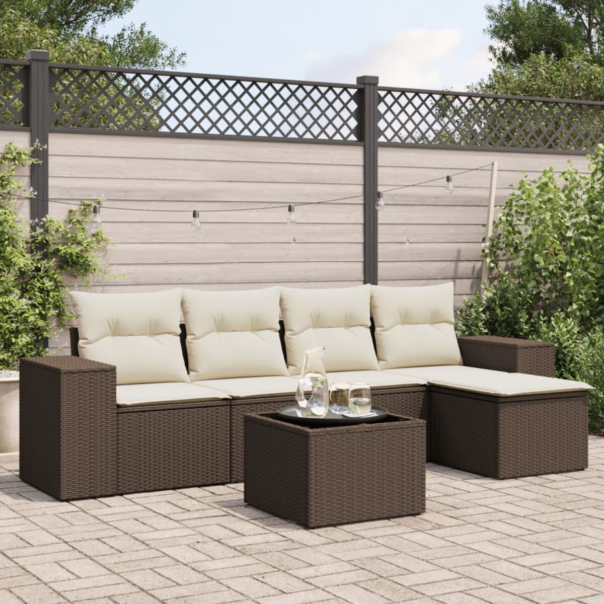 Braunes Korbmöbel-Set für die Terrasse mit cremefarbenen Kissen, Tisch und Getränken. Das Set steht auf einer Terrasse, Grün im Hintergrund.