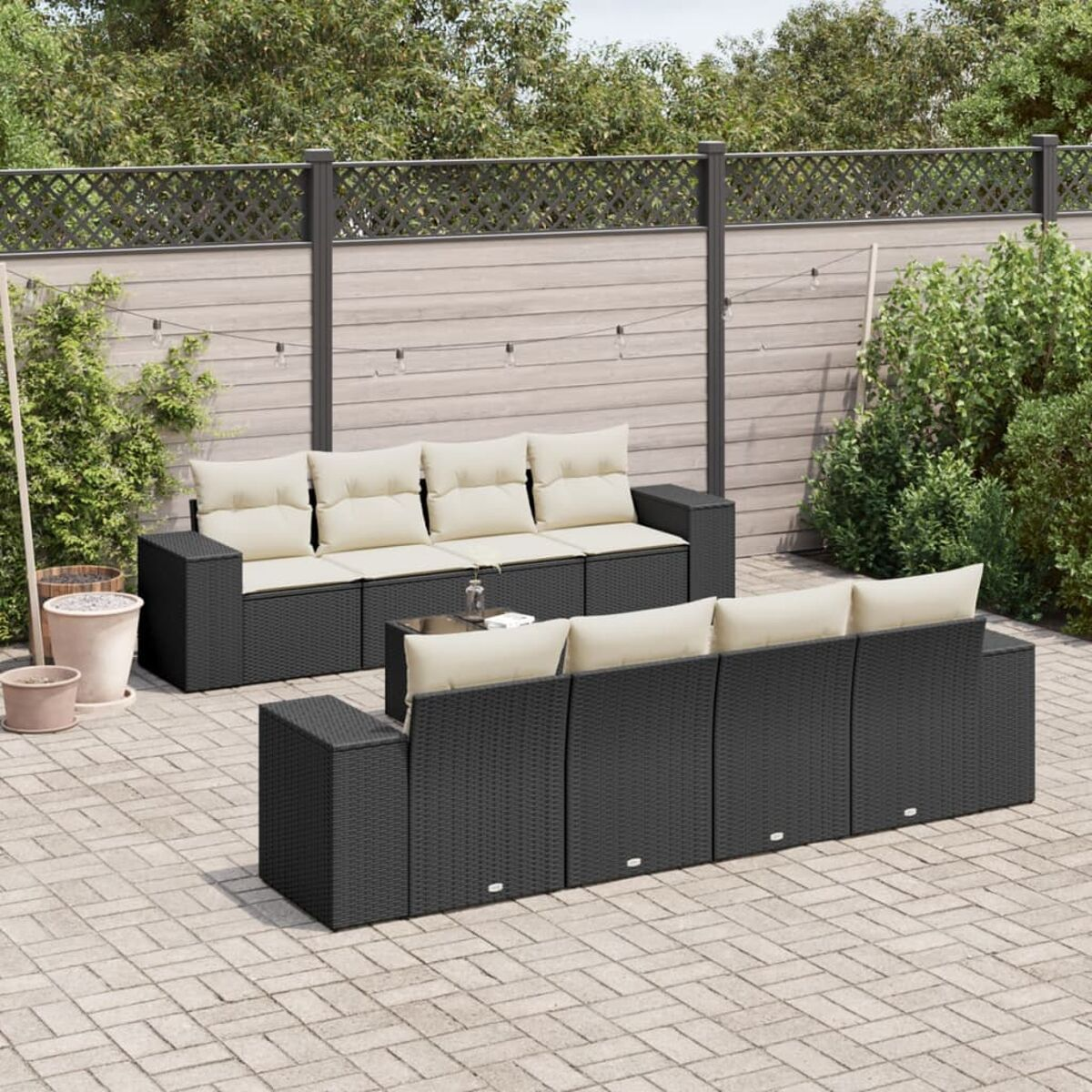 Schwarze Outdoor-Möbel aus Korbgeflecht auf einer Backstein-Terrasse. Cremefarbene Kissen, Zaun im Hintergrund.